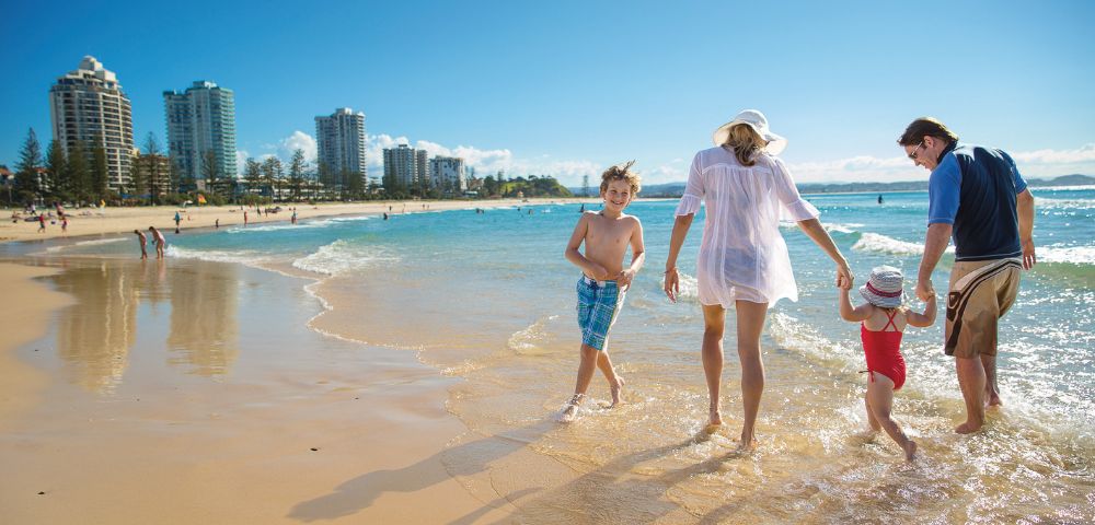 A family walks along a beach towards the ocean with city high-rises in the background.