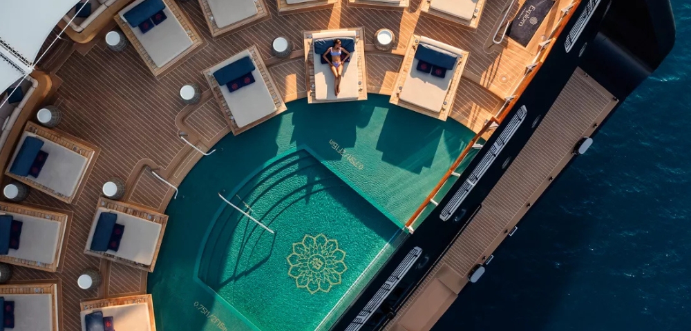 An aerial view of a luxury yacht deck featuring a curved swimming pool with a gold mandala design, surrounded by numerous lounge chairs and a person.