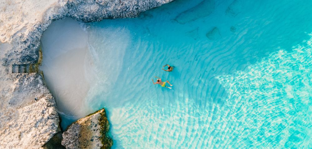 Aerial view of two people swimming in a shallow, crystal-clear turquoise lagoon surrounded by light-colored rocky cliffs and a small sandy beach.