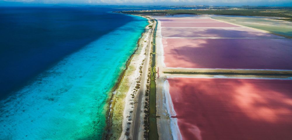 Aerial view of a narrow strip of land separating a vibrant turquoise sea from a series of large, rectangular pink salt ponds.