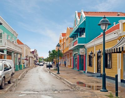 A narrow street lined with colorful Dutch colonial style buildings under a bright blue sky, with parked cars and a few pedestrians visible.