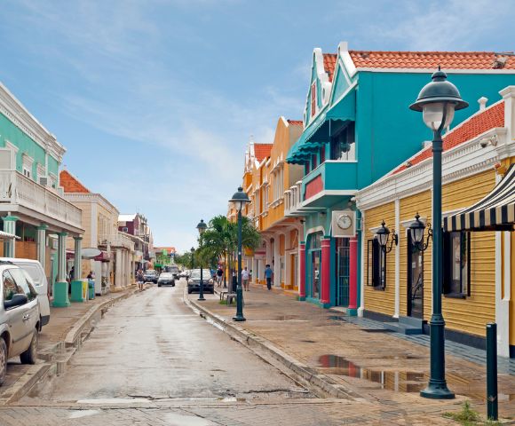 A narrow street lined with colorful Dutch colonial style buildings under a bright blue sky, with parked cars and a few pedestrians visible.