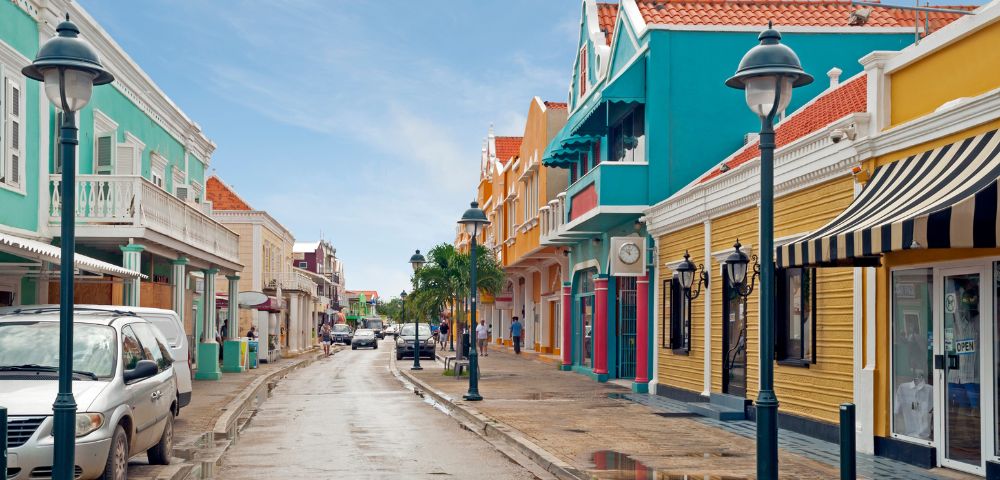 A narrow street lined with colorful Dutch colonial style buildings under a bright blue sky, with parked cars and a few pedestrians visible.