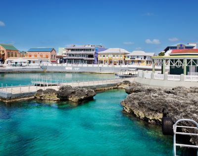 A panoramic view of a vibrant harbor with clear turquoise water, rocky outcrops, and a line of colorful buildings along the waterfront under a bright blue sky.