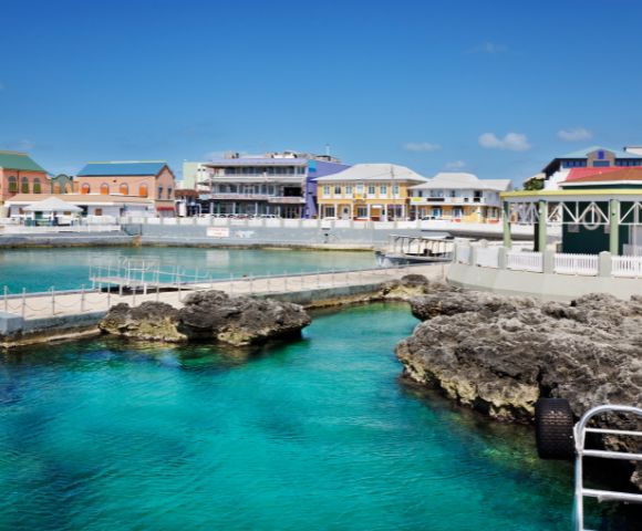 A panoramic view of a vibrant harbor with clear turquoise water, rocky outcrops, and a line of colorful buildings along the waterfront under a bright blue sky.