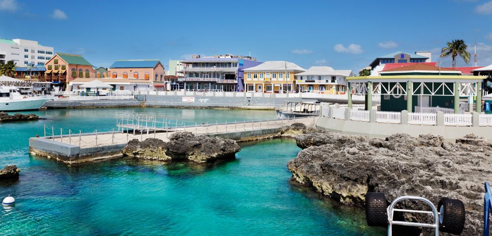 A panoramic view of a vibrant harbor with clear turquoise water, rocky outcrops, and a line of colorful buildings along the waterfront under a bright blue sky.
