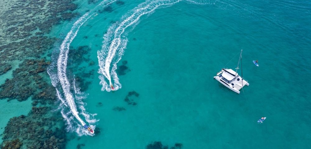 An aerial view of a turquoise sea with two jet skis creating wakes and a sailing boat anchored nearby.