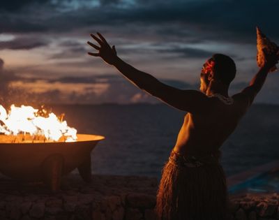 A person performing a traditional dance by a fire at dusk, with the ocean in the background.