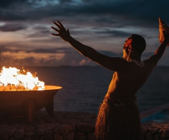 A person performing a traditional dance by a fire at dusk, with the ocean in the background.