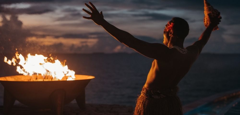 A person performing a traditional dance by a fire at dusk, with the ocean in the background.
