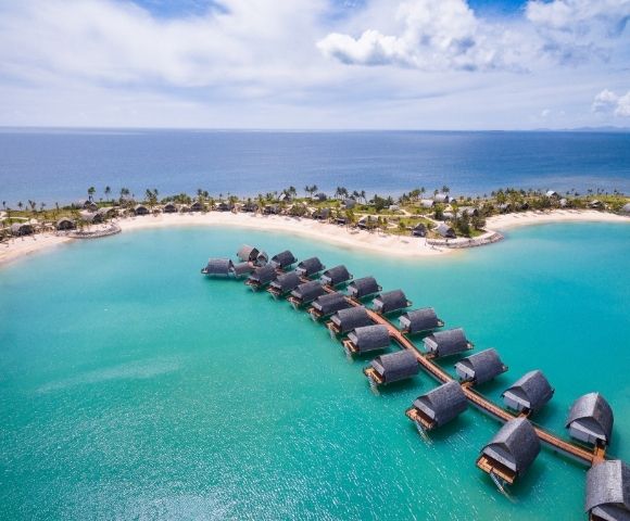 Aerial view of overwater bungalows in a turquoise lagoon near a sandy beach, with an island landscape in the background.