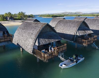 Overwater bungalows with thatched roofs line a calm turquoise bay, with a small boat navigating the water in the foreground and lush green islands in the background under a clear blue sky.