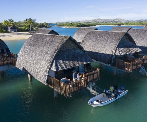 Overwater bungalows with thatched roofs line a calm turquoise bay, with a small boat navigating the water in the foreground and lush green islands in the background under a clear blue sky.