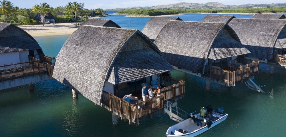 Overwater bungalows with thatched roofs line a calm turquoise bay, with a small boat navigating the water in the foreground and lush green islands in the background under a clear blue sky.