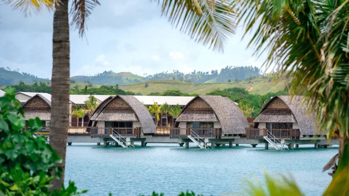 Row of overwater bungalows with thatched roofs on stilts over turquoise water, framed by palm fronds and lush green hills in the background.