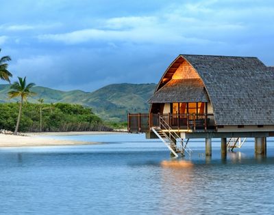 A peaceful beach scene featuring a wooden bungalow on stilts by the water, surrounded by palm trees and lush greenery, with mountains in the background under a cloudy sky.