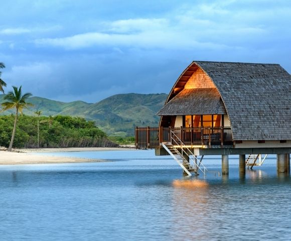 A peaceful beach scene featuring a wooden bungalow on stilts by the water, surrounded by palm trees and lush greenery, with mountains in the background under a cloudy sky.