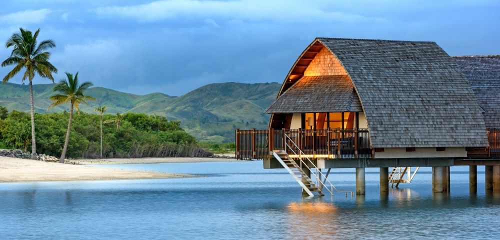 A peaceful beach scene featuring a wooden bungalow on stilts by the water, surrounded by palm trees and lush greenery, with mountains in the background under a cloudy sky.