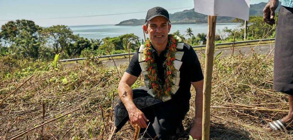 Man wearing a floral lei and black cap squats in a grassy area near the ocean with hills in the background under clear skies.