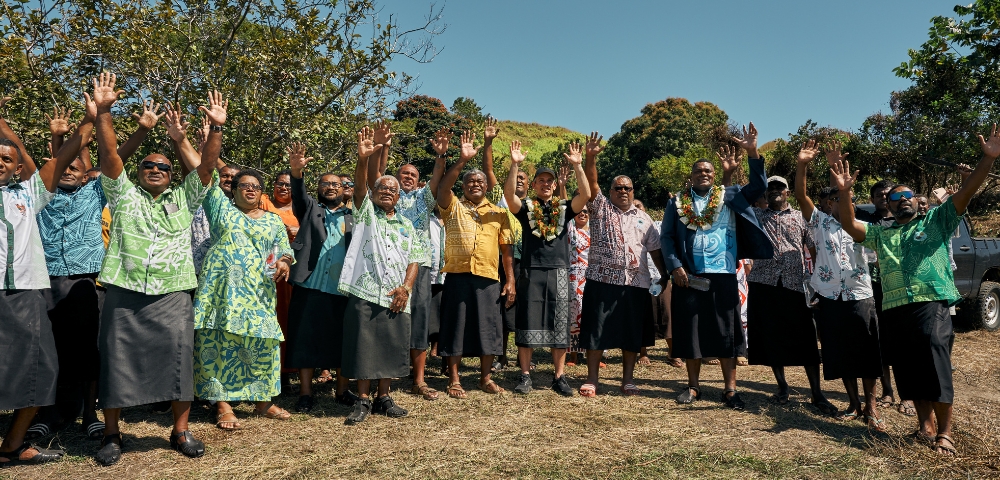 Group of people wearing traditional Pacific Island attire, standing outdoors on grass, smiling and waving hands in unison.