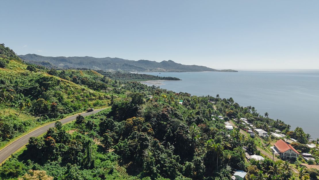 Coastal landscape with lush green hills, a winding road, scattered houses, and calm ocean waters under a clear sky.