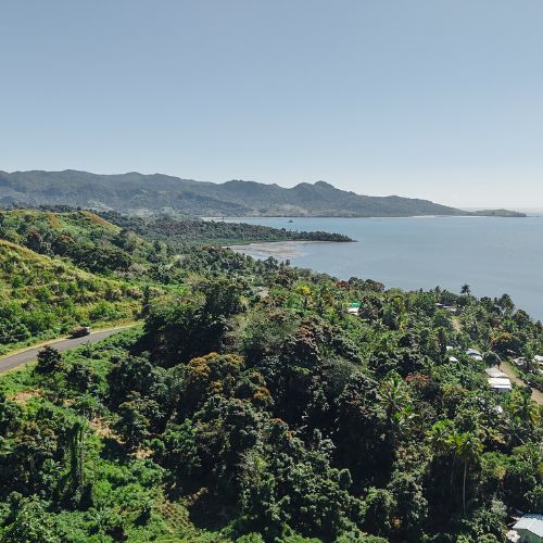 Coastal landscape with lush green hills, a winding road, scattered houses, and calm ocean waters under a clear sky.