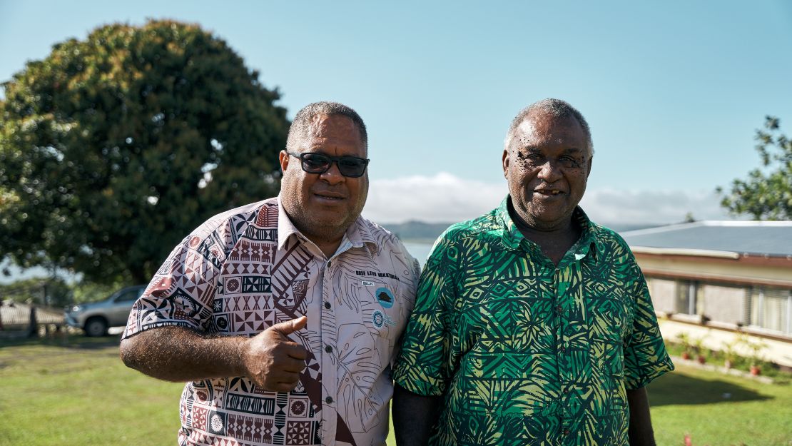 Two men in patterned shirts standing outdoors on a sunny day with a tree, car, and building in the background.