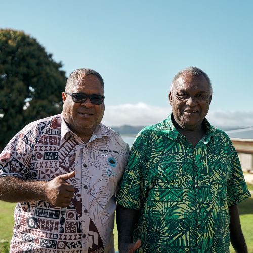 Two men in patterned shirts standing outdoors on a sunny day with a tree, car, and building in the background.