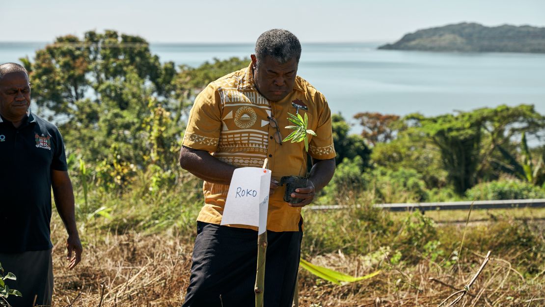 Man in a patterned yellow shirt holds a small plant next to a labeled stake in a grassy area with water and hills in the background.
