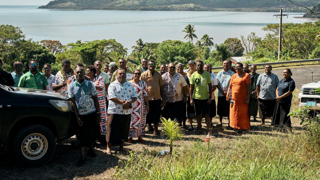 A group of people stand outdoors on a sunny day, with lush greenery and a tranquil body of water in the background. Everyone appears relaxed and gathered for a group photo.