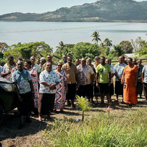 A group of people stand outdoors on a sunny day, with lush greenery and a tranquil body of water in the background. Everyone appears relaxed and gathered for a group photo.
