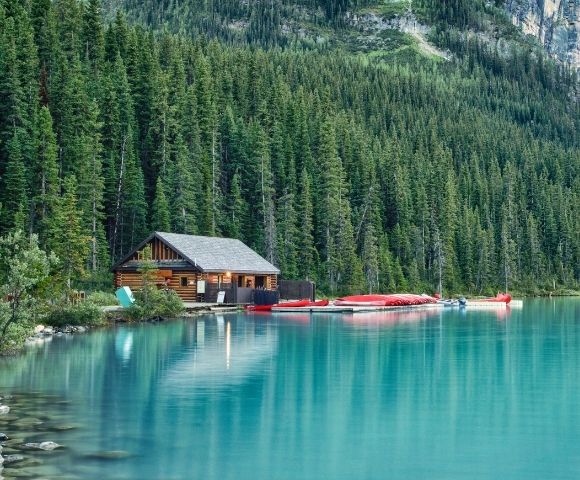 A serene lake surrounded by tall evergreen trees, featuring a wooden cabin and colorful canoes lined up at the water's edge.