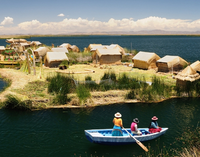 Floating reed island with thatched huts beside a deep-blue lake; three people paddle a small boat under a bright, peaceful sky.