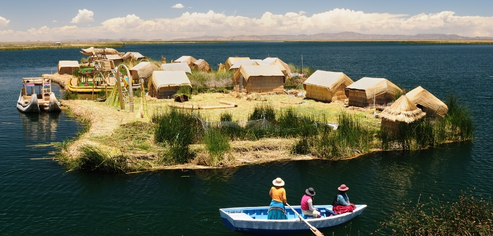 Floating reed island with thatched huts beside a deep-blue lake; three people paddle a small boat under a bright, peaceful sky.