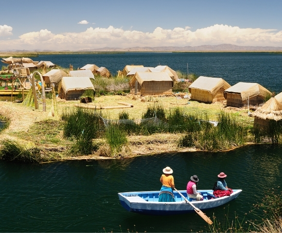 Floating reed island with thatched huts beside a deep-blue lake; three people paddle a small boat under a bright, peaceful sky.