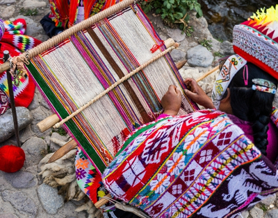 Indigenous Andean artisan in vibrant traditional clothing weaves a colorful patterned textile on a backstrap loom beside a rocky stream.
