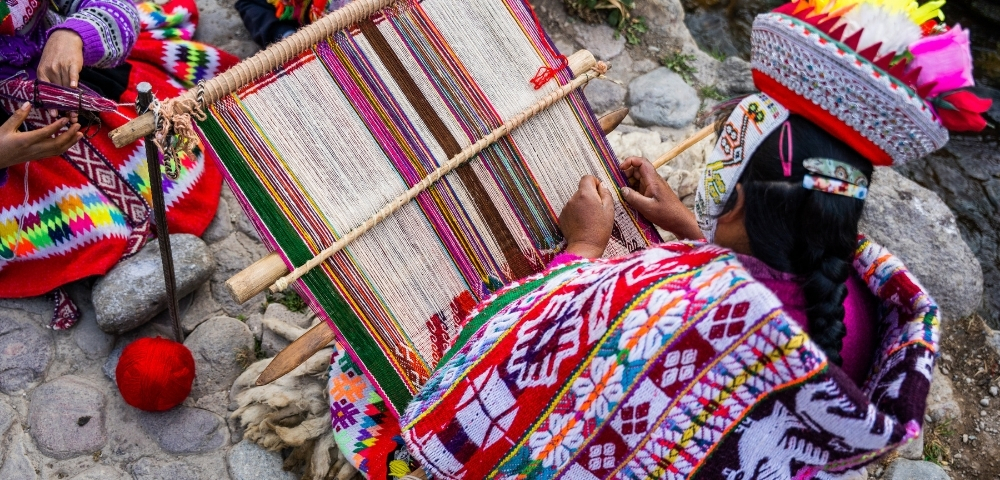 Indigenous Andean artisan in vibrant traditional clothing weaves a colorful patterned textile on a backstrap loom beside a rocky stream.