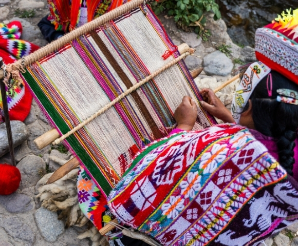 Indigenous Andean artisan in vibrant traditional clothing weaves a colorful patterned textile on a backstrap loom beside a rocky stream.