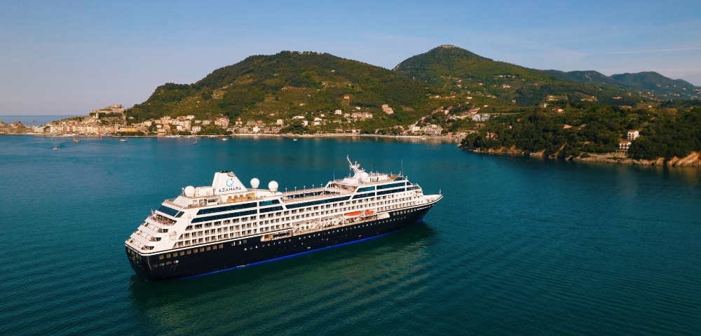 A large cruise ship sails in calm, blue waters near a lush, green coastline. The background features rolling hills and a small coastal town under a clear sky.