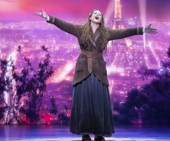 A woman with long brown hair and a brown braided jacket over a long blue skirt with a brown belt stands with her arms outstretched and head tilted back, singing on a stage with a vibrant purple and pink backdrop depicting the Eiffel Tower and a city skyline at night.