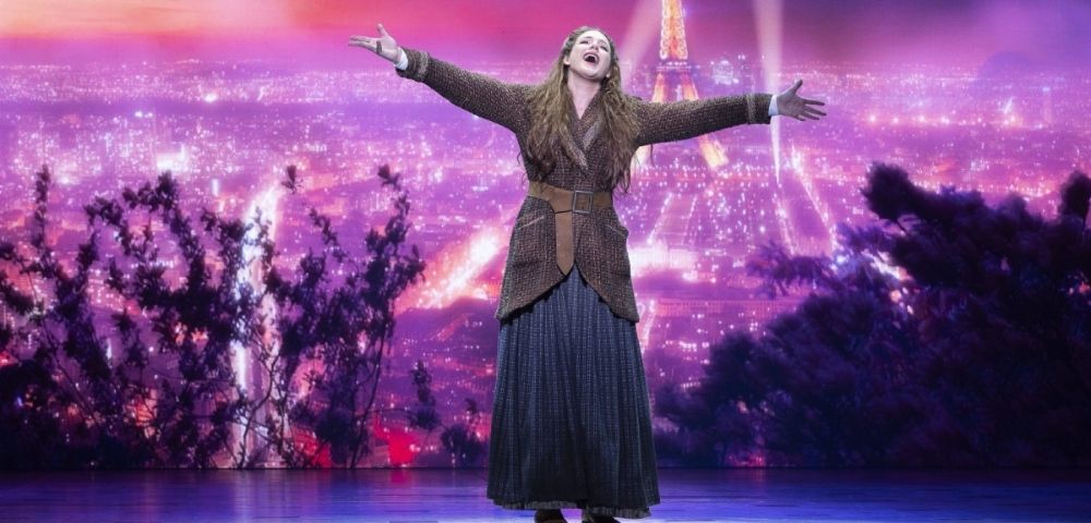 A woman with long brown hair and a brown braided jacket over a long blue skirt with a brown belt stands with her arms outstretched and head tilted back, singing on a stage with a vibrant purple and pink backdrop depicting the Eiffel Tower and a city skyline at night.
