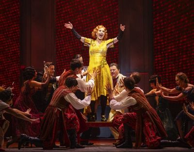 A woman in a bright yellow dress stands on a raised platform on a stage, with her arms outstretched and a wide smile on her face. She is surrounded by a group of performers in red and brown costumes. The background is a dark red, textured wall.