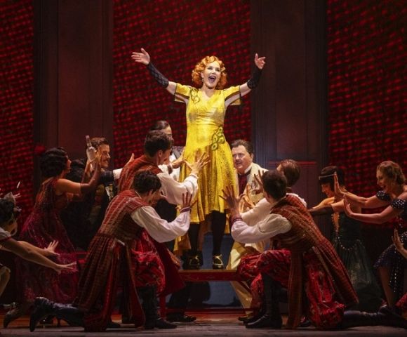 A woman in a bright yellow dress stands on a raised platform on a stage, with her arms outstretched and a wide smile on her face. She is surrounded by a group of performers in red and brown costumes. The background is a dark red, textured wall.