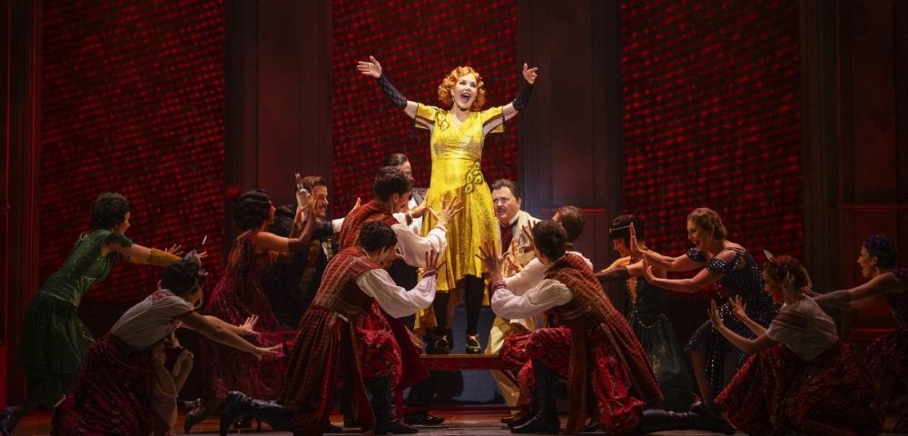A woman in a bright yellow dress stands on a raised platform on a stage, with her arms outstretched and a wide smile on her face. She is surrounded by a group of performers in red and brown costumes. The background is a dark red, textured wall.