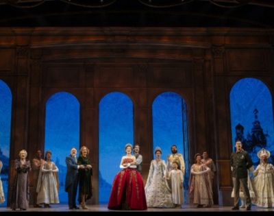 A stage performance with a group of actors in elaborate historical costumes, standing in front of a backdrop featuring arched windows showing iconic cityscapes, including the Eiffel Tower and Russian architecture, under a deep blue sky.