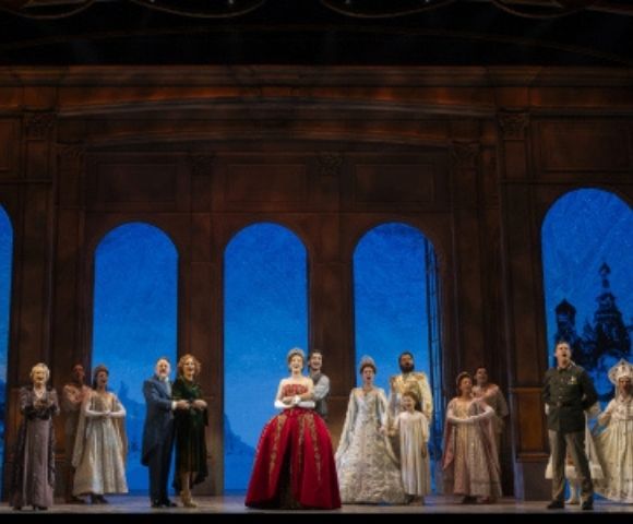 A stage performance with a group of actors in elaborate historical costumes, standing in front of a backdrop featuring arched windows showing iconic cityscapes, including the Eiffel Tower and Russian architecture, under a deep blue sky.