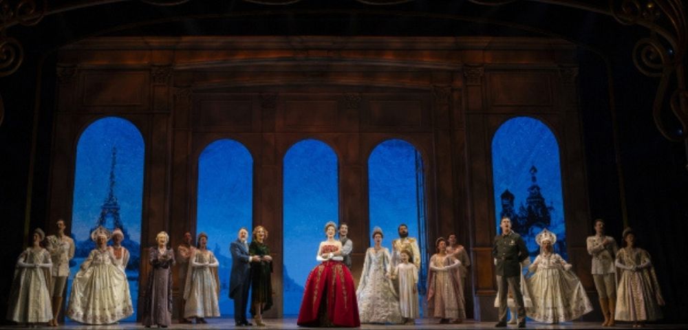 A stage performance with a group of actors in elaborate historical costumes, standing in front of a backdrop featuring arched windows showing iconic cityscapes, including the Eiffel Tower and Russian architecture, under a deep blue sky.