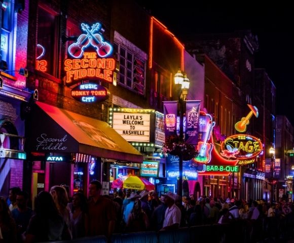 A vibrant Nashville street at night, featuring bright neon signs for The Second Fiddle and The Stage, crowded with people walking under lights.