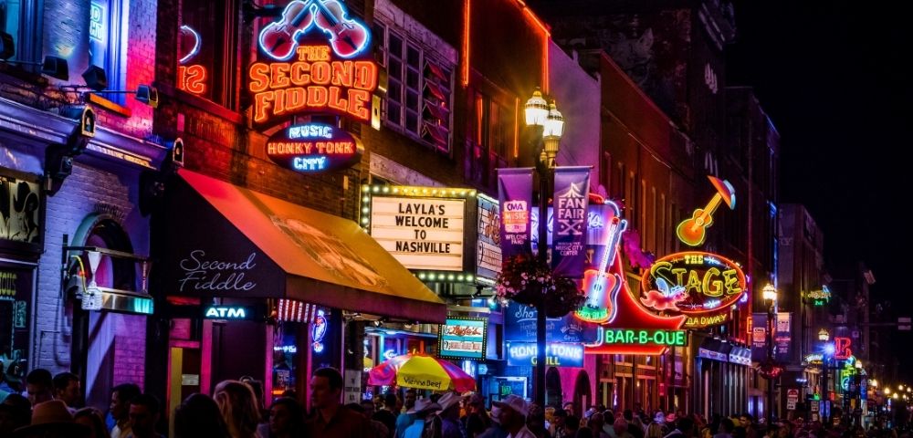 A vibrant Nashville street at night, featuring bright neon signs for The Second Fiddle and The Stage, crowded with people walking under lights.