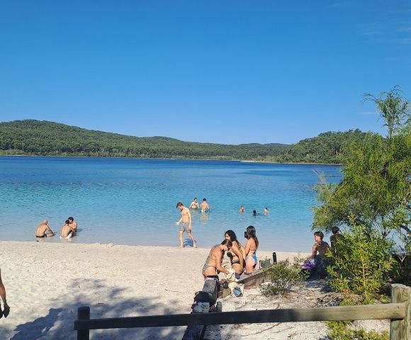 A beach scene with people swimming and relaxing near clear blue water and green hills in the background.